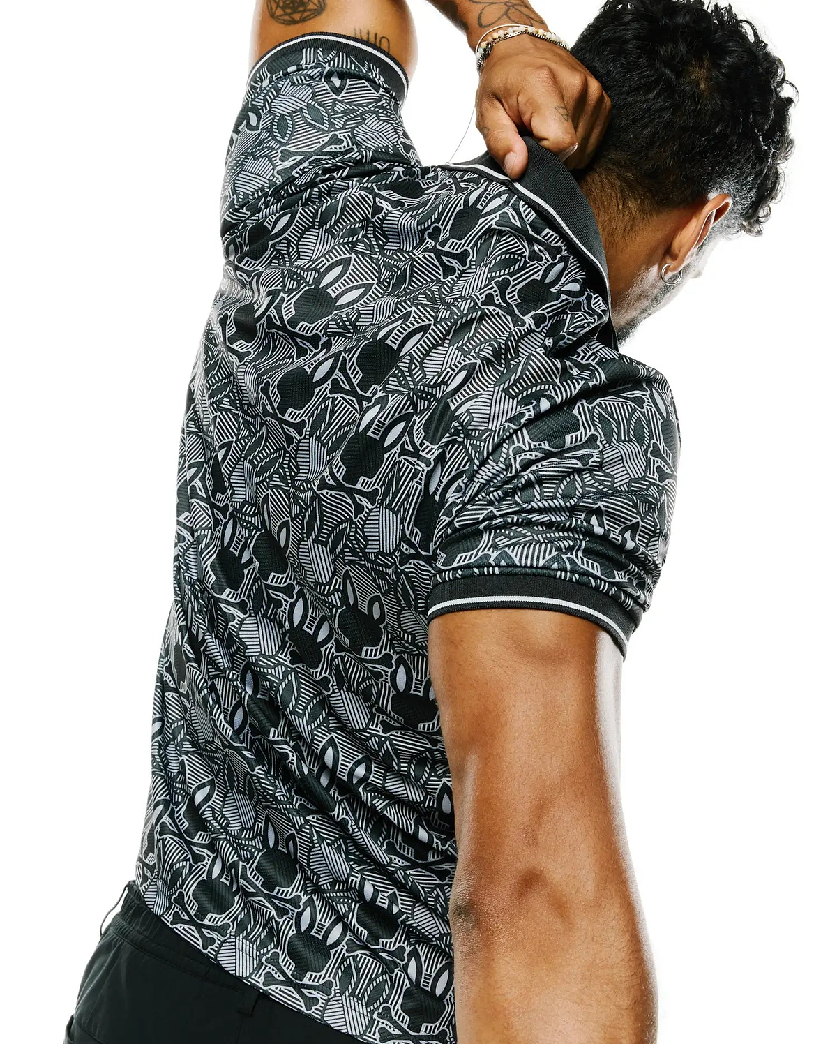 Mens Reeves All Over Bunny Sport Polo, a black and white performance jersey polo with a classic fit, featuring an intricate all-over Psycho Bunny skull and geometric pattern, with white striped collar and sleeve cuffs.