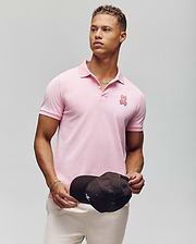 A young man with short curly hair wears the Psycho Bunny MENS PATRICK MENS POLO - MT0100095 in light pink 100% cotton, featuring a small bunny logo. He stands against a plain light background, holding a black cap and looking to the side. (1)