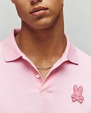 A person wears the Psycho Bunny MENS PATRICK MENS POLO - MT0100095 in light pink, featuring the signature bunny and crossbones logo, styled with a thin silver chain. The image is cropped to show only the lower face and upper torso. (2)