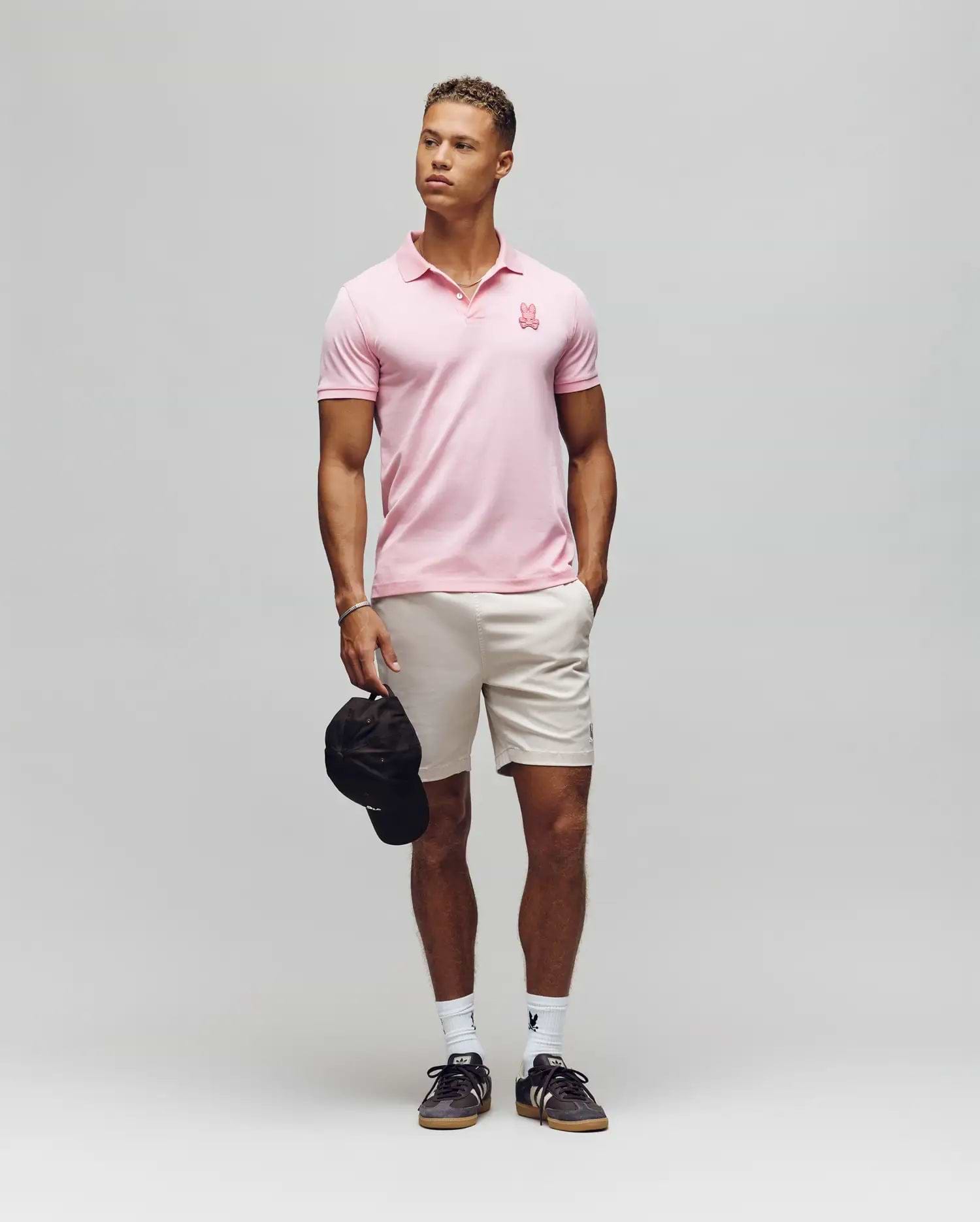 A man poses against a plain background wearing the Psycho Bunny MENS PATRICK MENS POLO - MT0100095 in light pink, beige shorts, white socks, and black sneakers. He holds a black cap in his left hand and looks slightly to his right. (3)