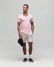 A man poses against a plain background wearing the Psycho Bunny MENS PATRICK MENS POLO - MT0100095 in light pink, beige shorts, white socks, and black sneakers. He holds a black cap in his left hand and looks slightly to his right. (3)
