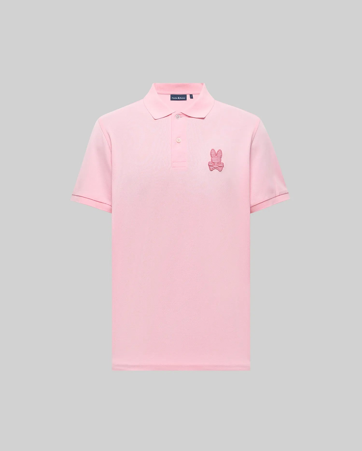 The Psycho Bunny MENS PATRICK POLO (MT0100095) is a light pink, 100% cotton knit short-sleeve shirt with a collar, button placket, and an embroidered pink bunny logo on the left chest, shown against a gray background. (4)