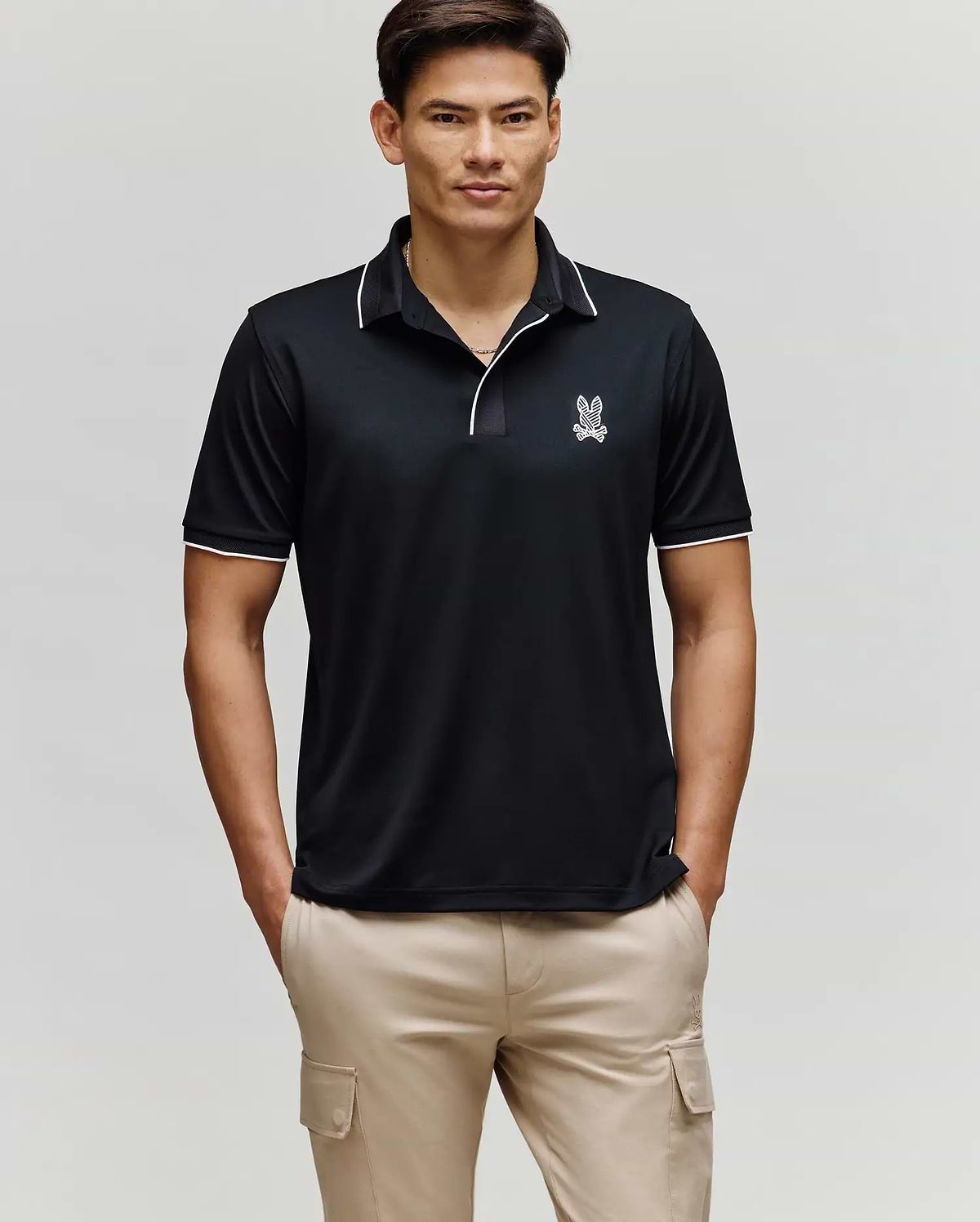A man stands facing forward wearing the Psycho Bunny MENS JAYDEN SPORT POLO - MT0100111 in black with white trim and a small bunny logo on the chest, paired with light beige cargo pants. He has his hands in his pockets and a neutral expression.