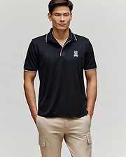 A man stands facing forward wearing the Psycho Bunny MENS JAYDEN SPORT POLO - MT0100111 in black with white trim and a small bunny logo on the chest, paired with light beige cargo pants. He has his hands in his pockets and a neutral expression.