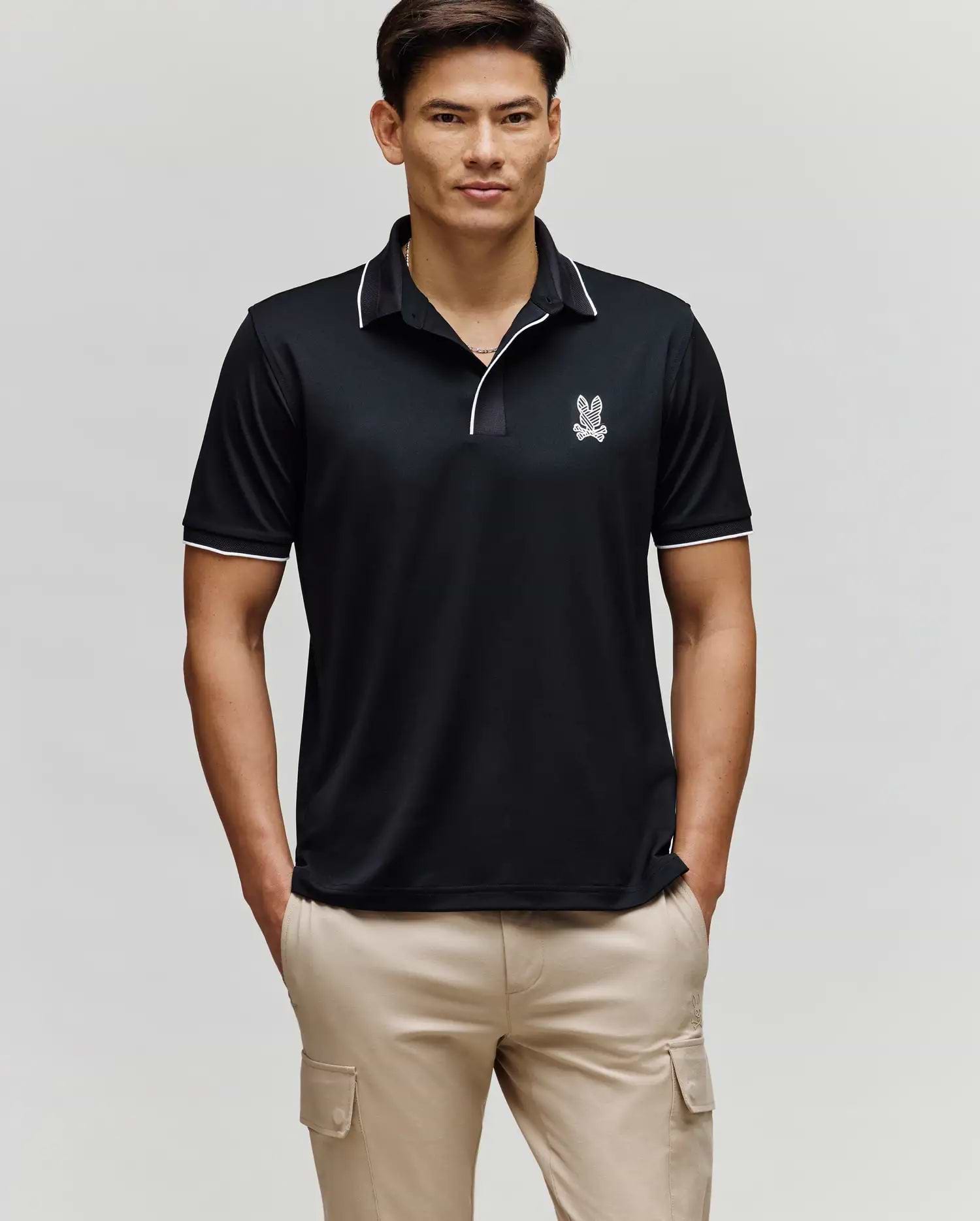 A man stands facing forward wearing the Psycho Bunny MENS JAYDEN SPORT POLO - MT0100111 in black with white trim and a small bunny logo on the chest, paired with light beige cargo pants. He has his hands in his pockets and a neutral expression.