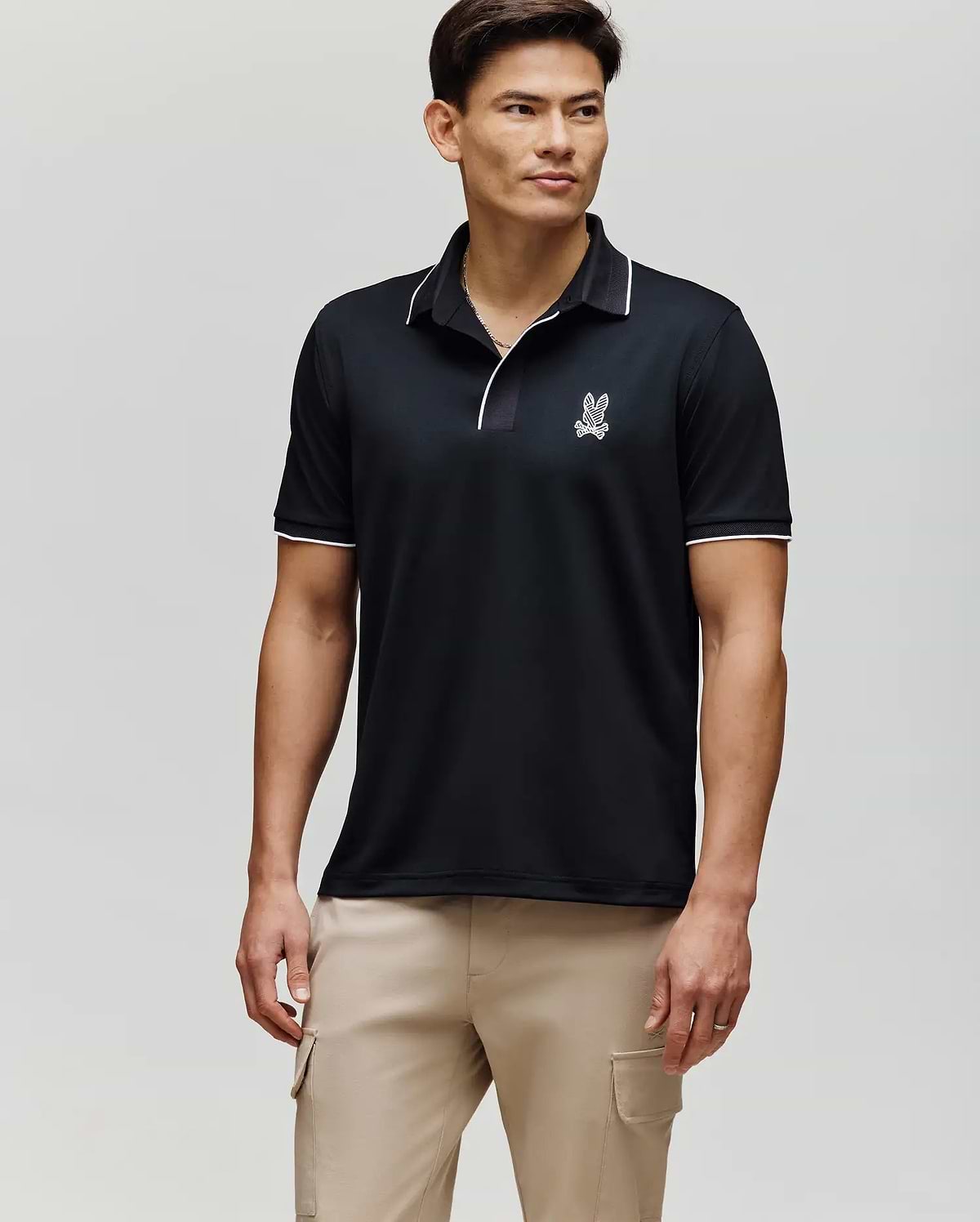 A man with short dark hair stands facing forward in a Psycho Bunny MENS JAYDEN SPORT POLO (MT0100111), featuring a black athletic fit with white trim and a small logo on the chest, paired with light beige cargo pants against a light gray background.