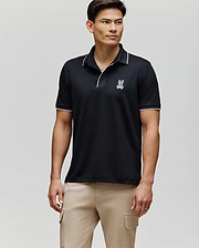 A man with short dark hair stands facing forward in a Psycho Bunny MENS JAYDEN SPORT POLO (MT0100111), featuring a black athletic fit with white trim and a small logo on the chest, paired with light beige cargo pants against a light gray background.