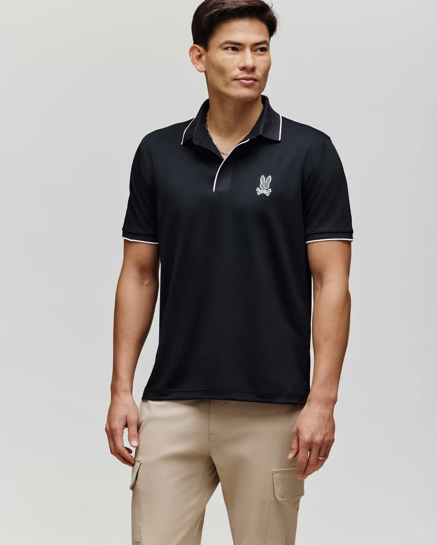 A man with short dark hair stands facing forward in a Psycho Bunny MENS JAYDEN SPORT POLO (MT0100111), featuring a black athletic fit with white trim and a small logo on the chest, paired with light beige cargo pants against a light gray background.