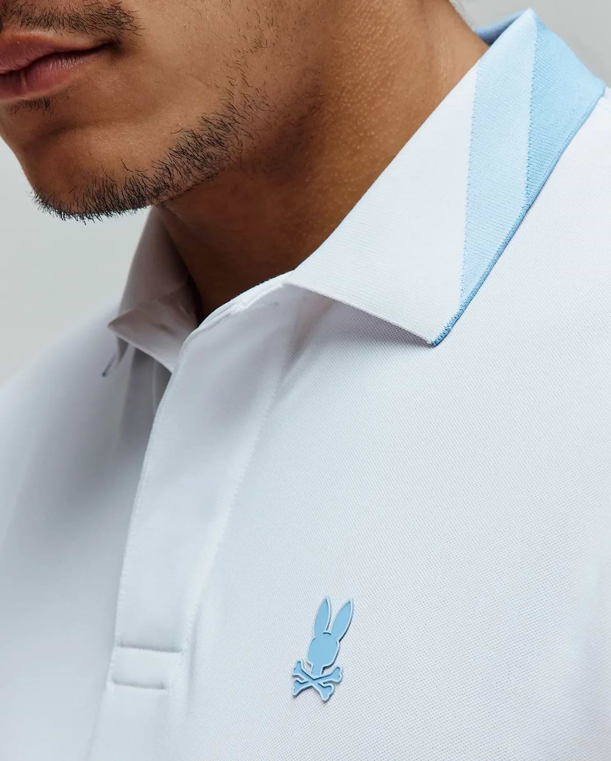 Mens Onyx Sport Polo, a crisp white pique knit polo shirt featuring a classic fit, a light blue striped collar, and a small, raised light blue Psycho Bunny skull logo on the left chest. (2)