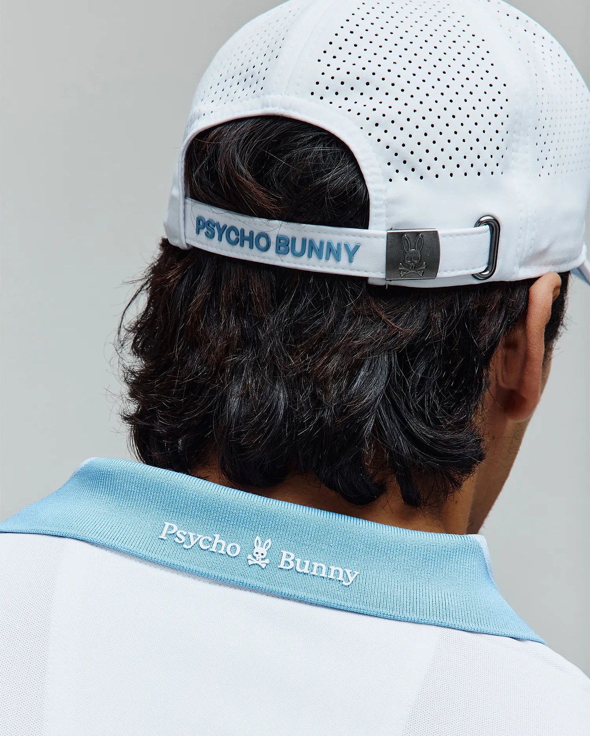 Mens Onyx Sport Polo in white pique with a light blue ribbed collar featuring white embroidered Psycho Bunny text and skull logo on the back, paired with a white perforated sport cap with light blue Psycho Bunny text and a metal bunny skull buckle. (3)
