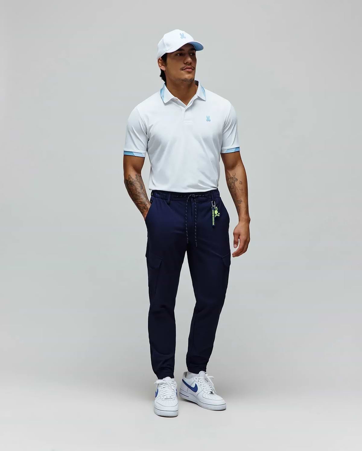 White Mens Onyx Sport Polo with light blue collar trim and cuffs, featuring a small embroidered Psycho Bunny logo, paired with navy cargo joggers with a neon green Psycho Bunny keychain, and a white cap with a light blue logo. (4)