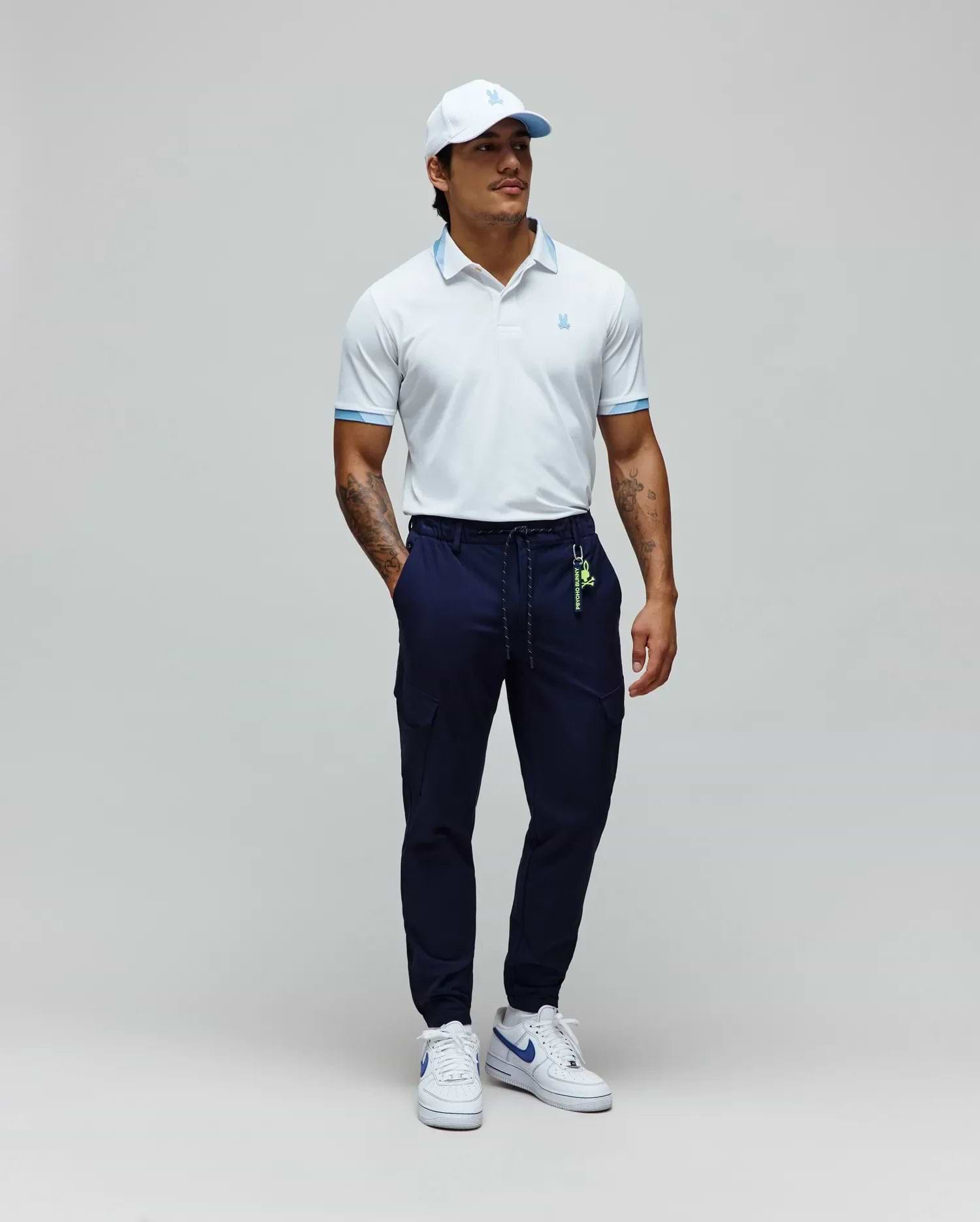 White Mens Onyx Sport Polo with light blue collar trim and cuffs, featuring a small embroidered Psycho Bunny logo, paired with navy cargo joggers with a neon green Psycho Bunny keychain, and a white cap with a light blue logo. (4)