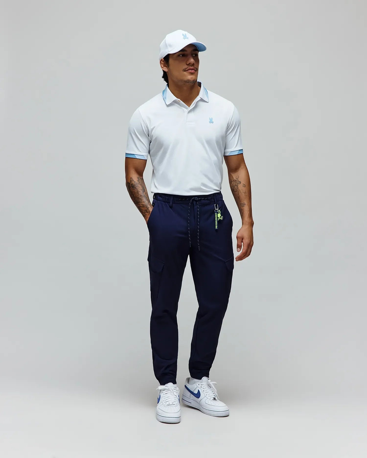 White Mens Onyx Sport Polo with light blue collar trim and cuffs, featuring a small embroidered Psycho Bunny logo, paired with navy cargo joggers with a neon green Psycho Bunny keychain, and a white cap with a light blue logo. (4)