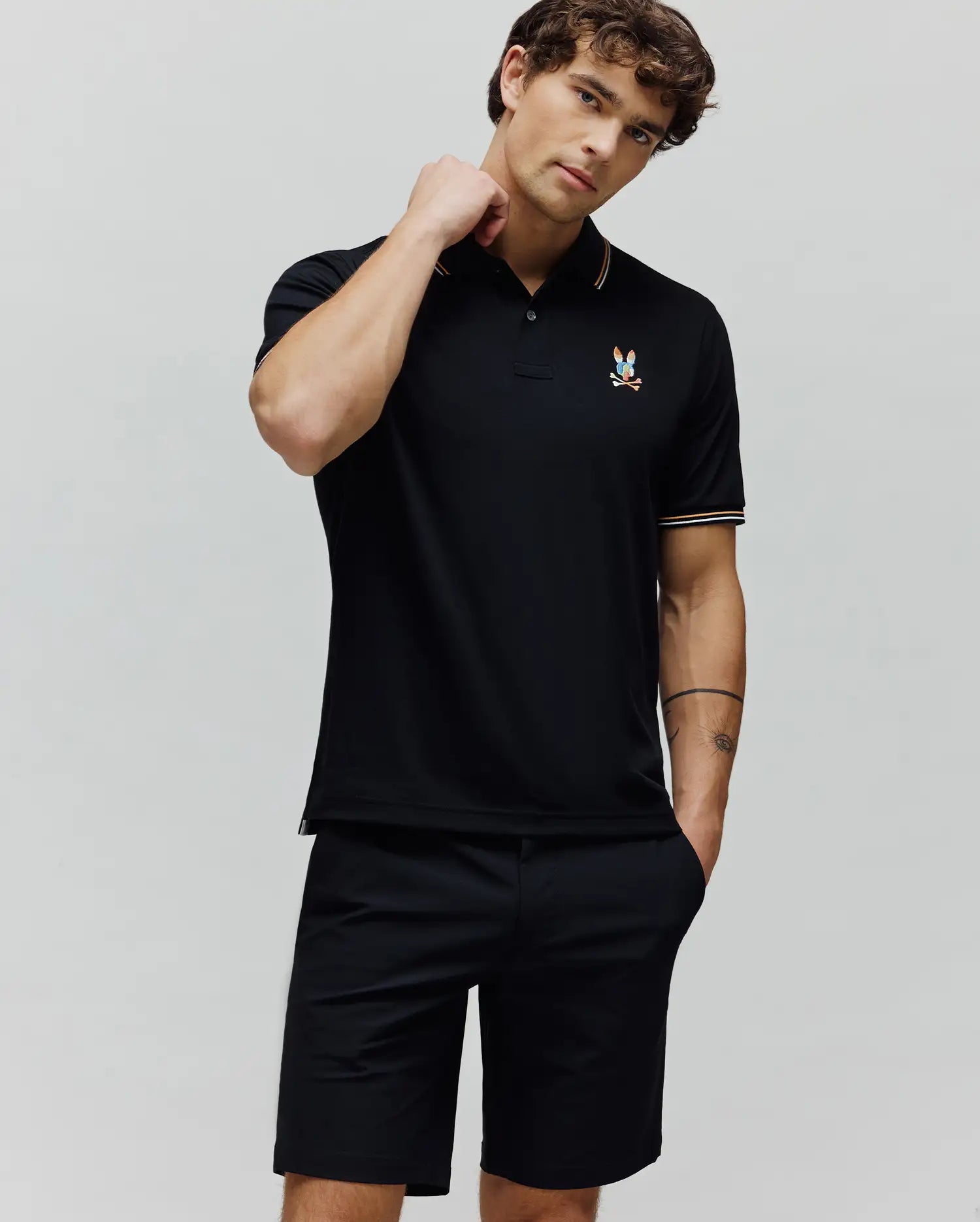 Black Mens Ara Pique Polo in a slim fit, featuring a pique texture, colorful embroidered Psycho Bunny skull logo on the left chest, and orange and white tipping stripes on the collar and sleeve cuffs.