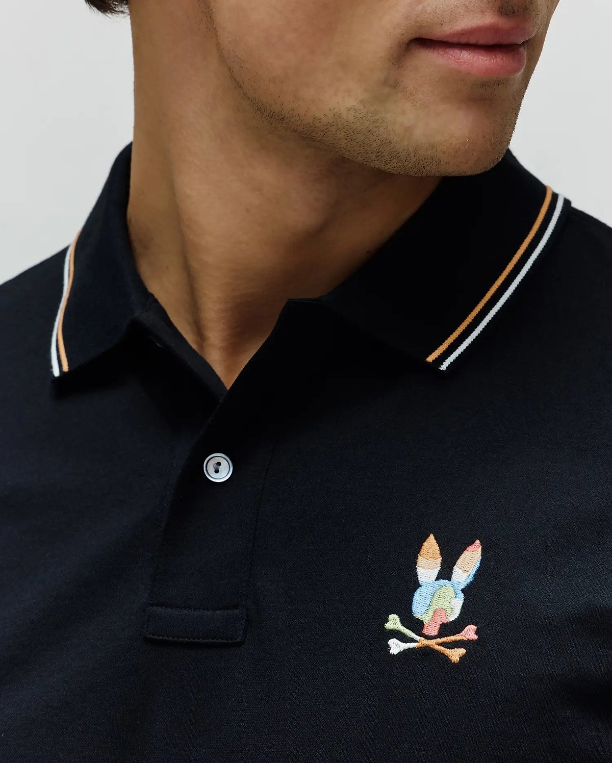 Black Mens Ara Pique Polo with white and orange tipping on the collar and a small, embroidered, multicolored Psycho Bunny skull and crossbones logo on the left chest.