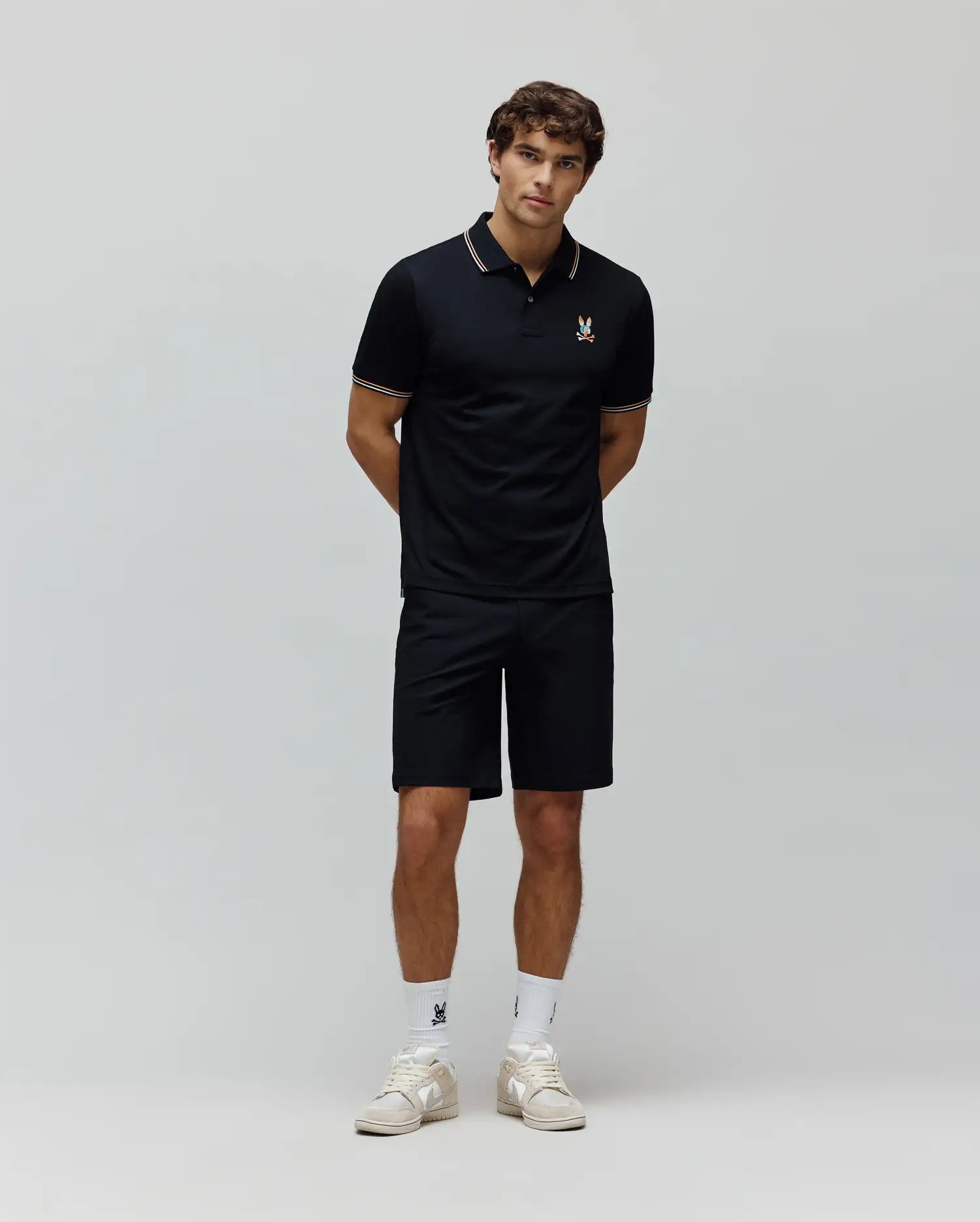 Mens Ara Pique Polo in navy, classic fit, with white and orange striped collar and cuffs, featuring a small, colorful embroidered Psycho Bunny skull logo on the left chest, paired with navy shorts.