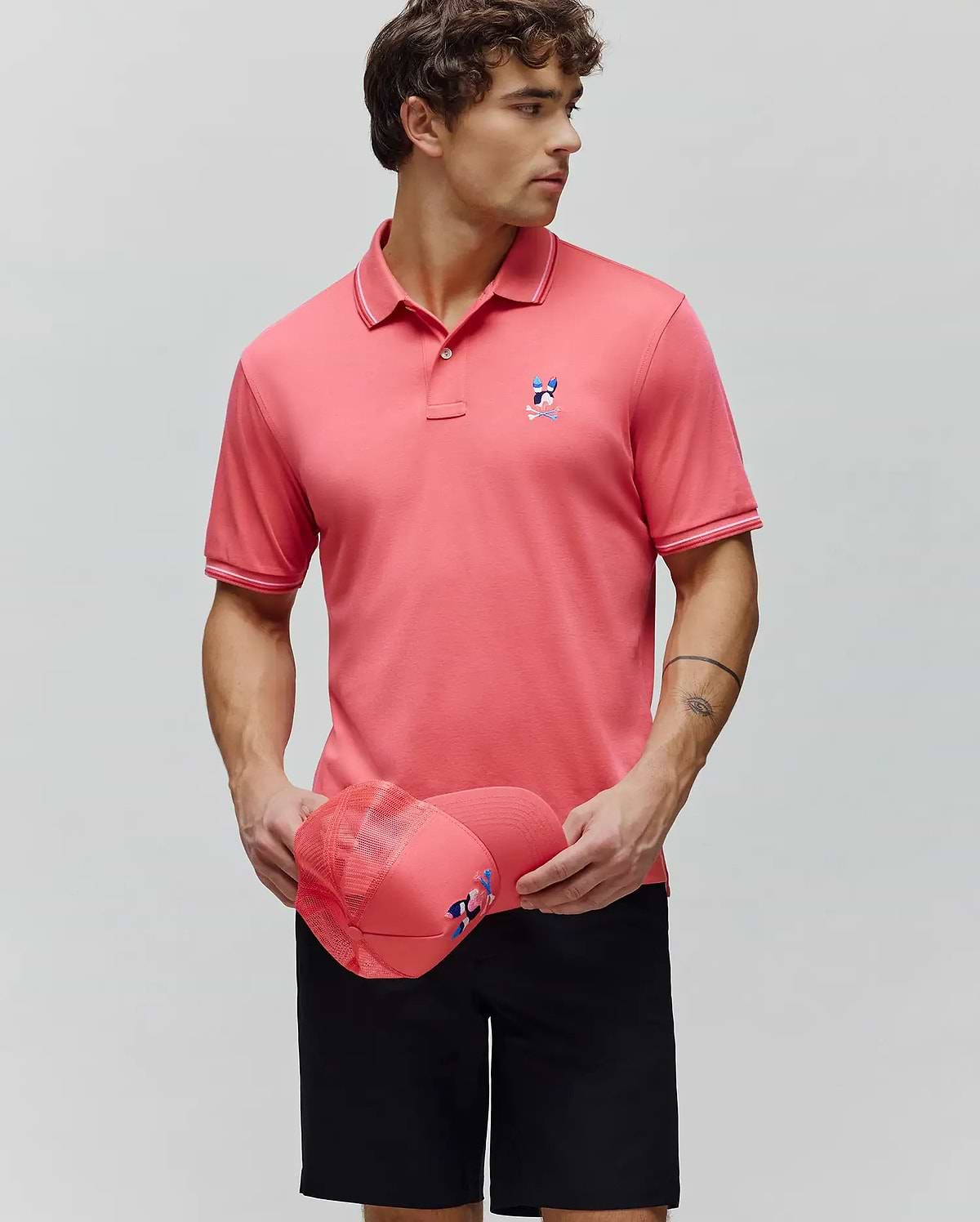 Mens Ara Pique Polo in vibrant coral pique knit, featuring a classic fit, white tipping on the collar and cuffs, and a colorful embroidered Psycho Bunny skull logo on the left chest. (1)