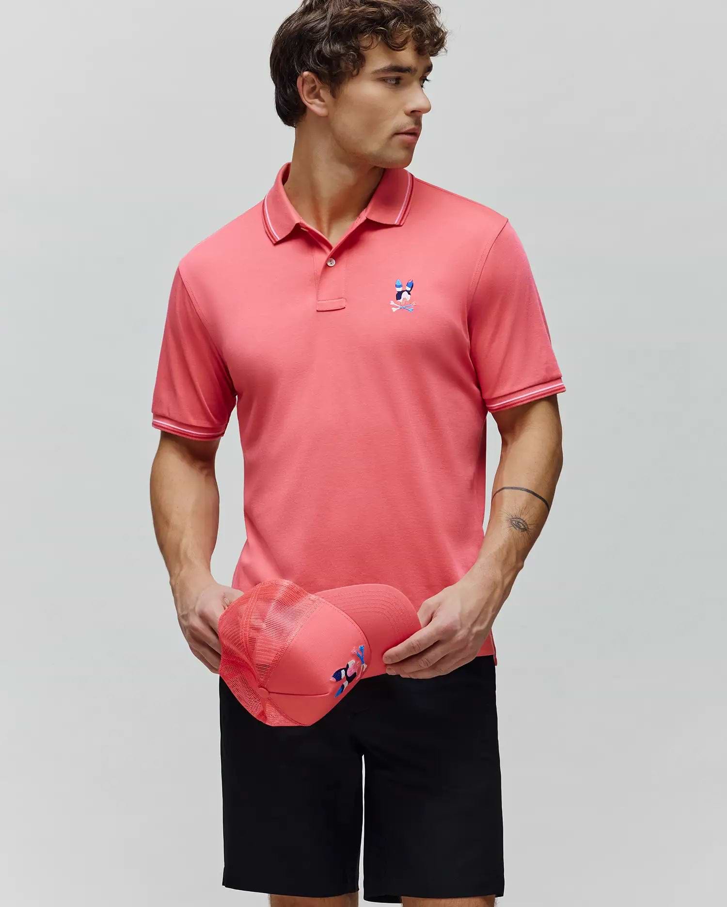 Mens Ara Pique Polo in vibrant coral pique knit, featuring a classic fit, white tipping on the collar and cuffs, and a colorful embroidered Psycho Bunny skull logo on the left chest. (1)