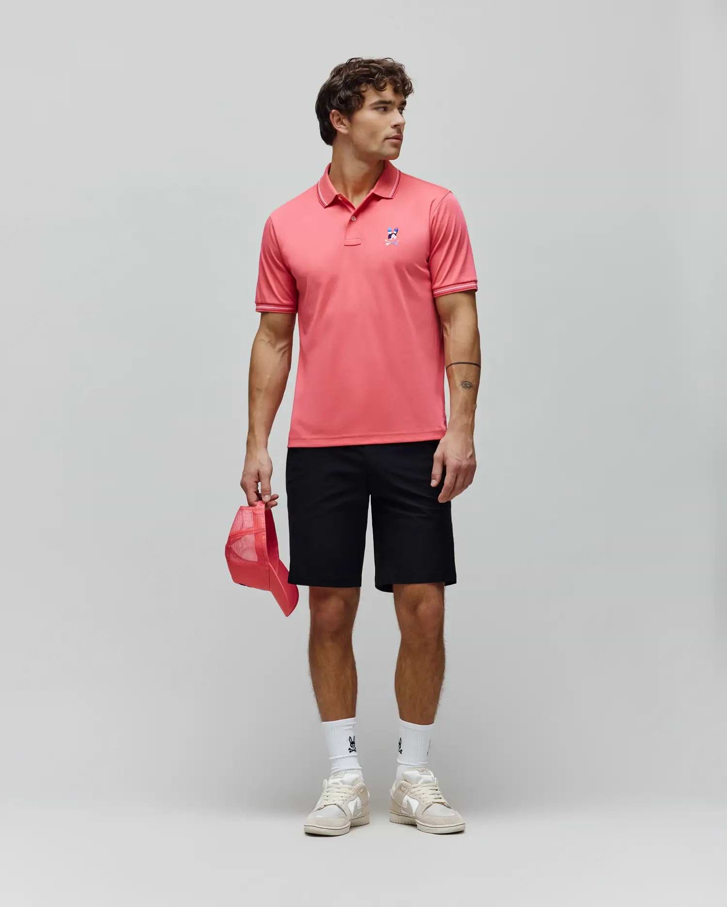 Mens Ara Pique Polo in coral, classic fit, with white tipping and a multi-color embroidered bunny skull logo on the left chest. Black shorts, white bunny skull socks, and beige sneakers. (4)