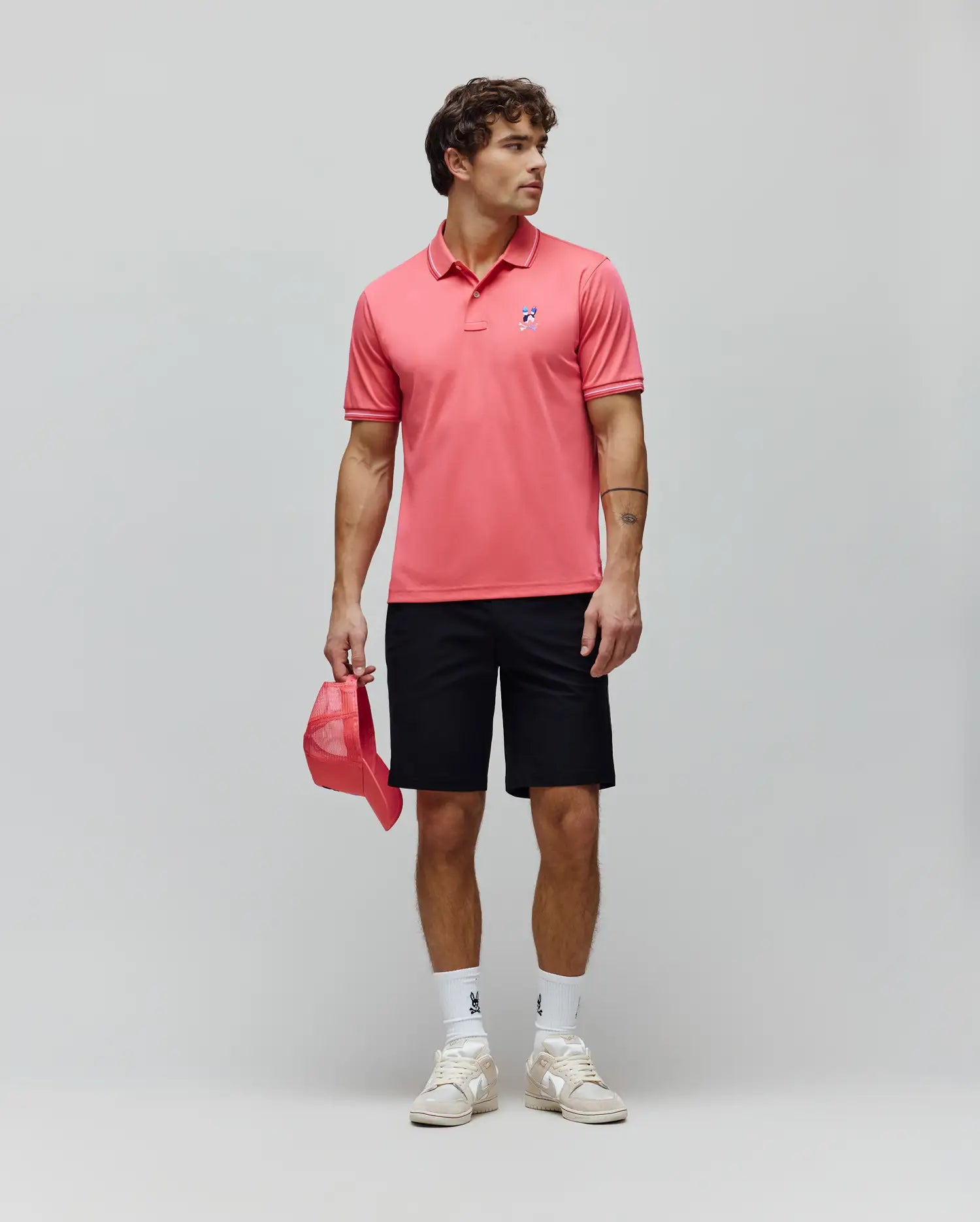 Mens Ara Pique Polo in coral, classic fit, with white tipping and a multi-color embroidered bunny skull logo on the left chest. Black shorts, white bunny skull socks, and beige sneakers. (4)