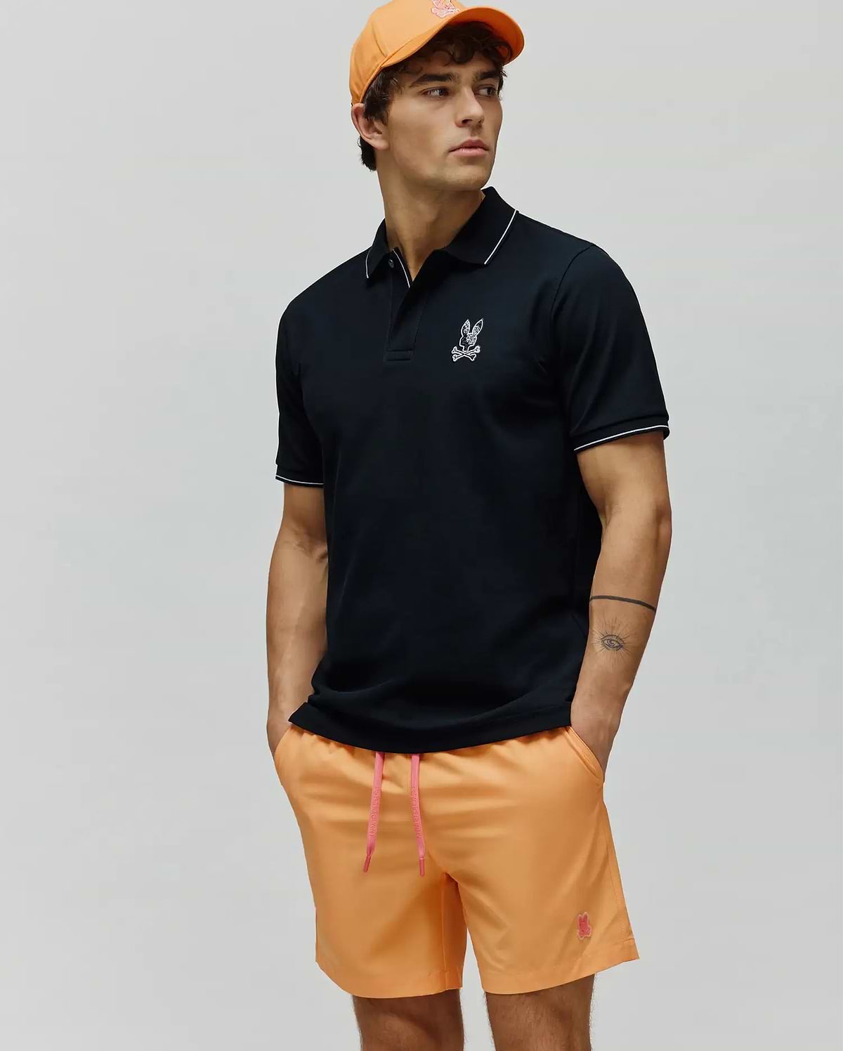 Mens Olivier Pique Polo in black pique with white tipping and embroidered Psycho Bunny skull logo on chest, paired with orange shorts featuring a pink drawstring and embroidered logo, plus an orange cap. (3)