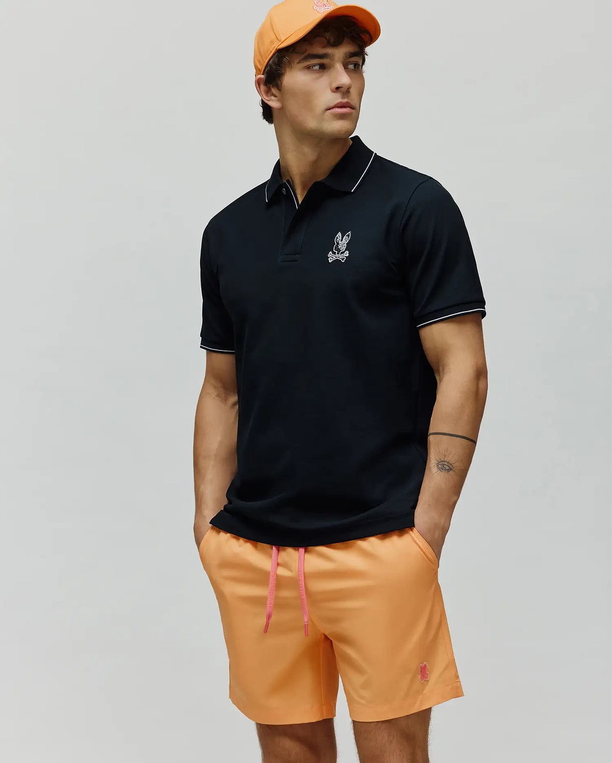 Mens Olivier Pique Polo in black pique with white tipping and embroidered Psycho Bunny skull logo on chest, paired with orange shorts featuring a pink drawstring and embroidered logo, plus an orange cap. (3)