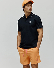 Mens Olivier Pique Polo in black pique with white tipping and embroidered Psycho Bunny skull logo on chest, paired with orange shorts featuring a pink drawstring and embroidered logo, plus an orange cap. (3)