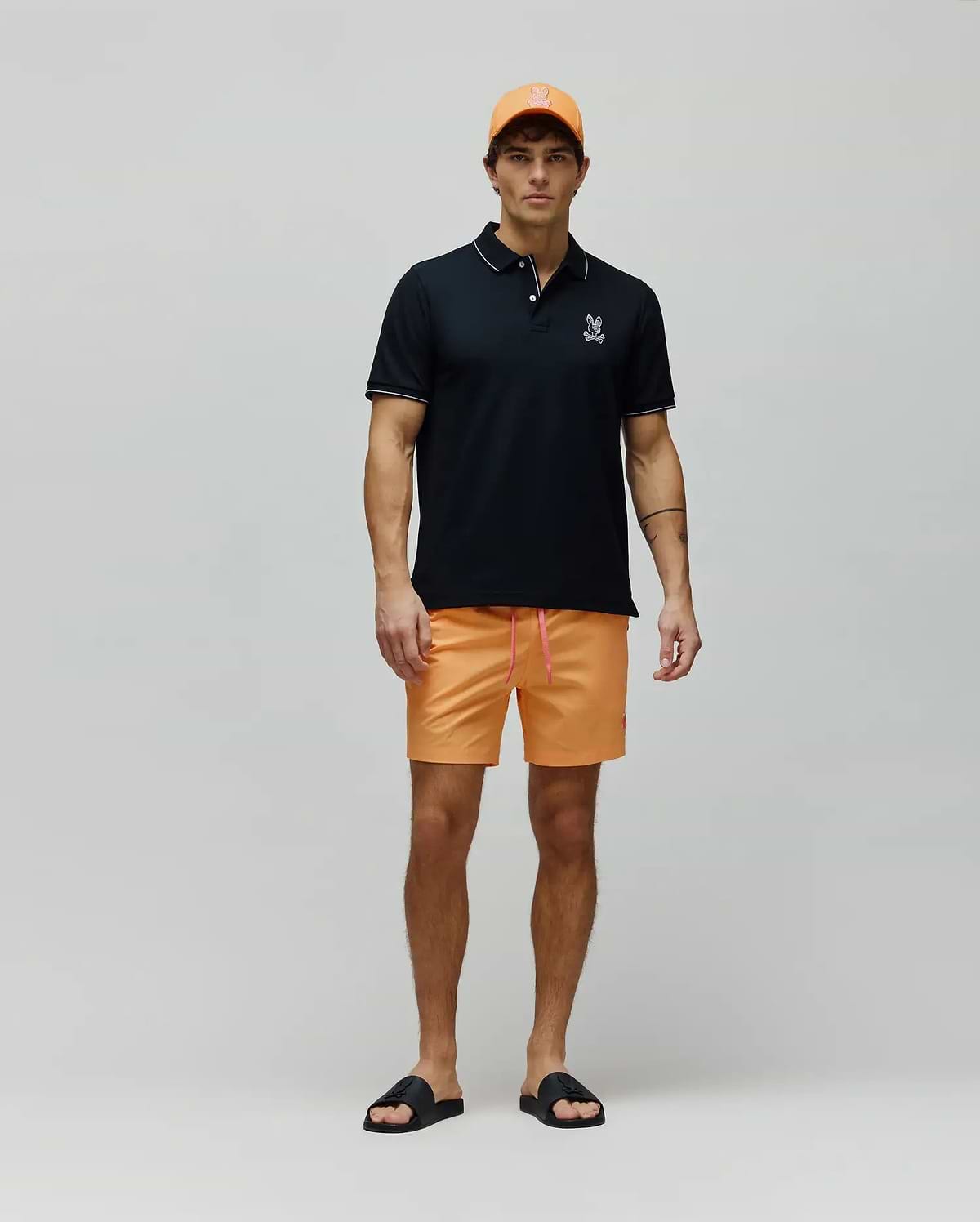 Mens Olivier Pique Polo in black with white trim and embroidered Psycho Bunny logo, paired with bright orange shorts featuring a pink drawstring and small bunny logo, an orange cap, and black logo slides. (4)
