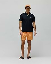 Mens Olivier Pique Polo in black with white trim and embroidered Psycho Bunny logo, paired with bright orange shorts featuring a pink drawstring and small bunny logo, an orange cap, and black logo slides. (4)