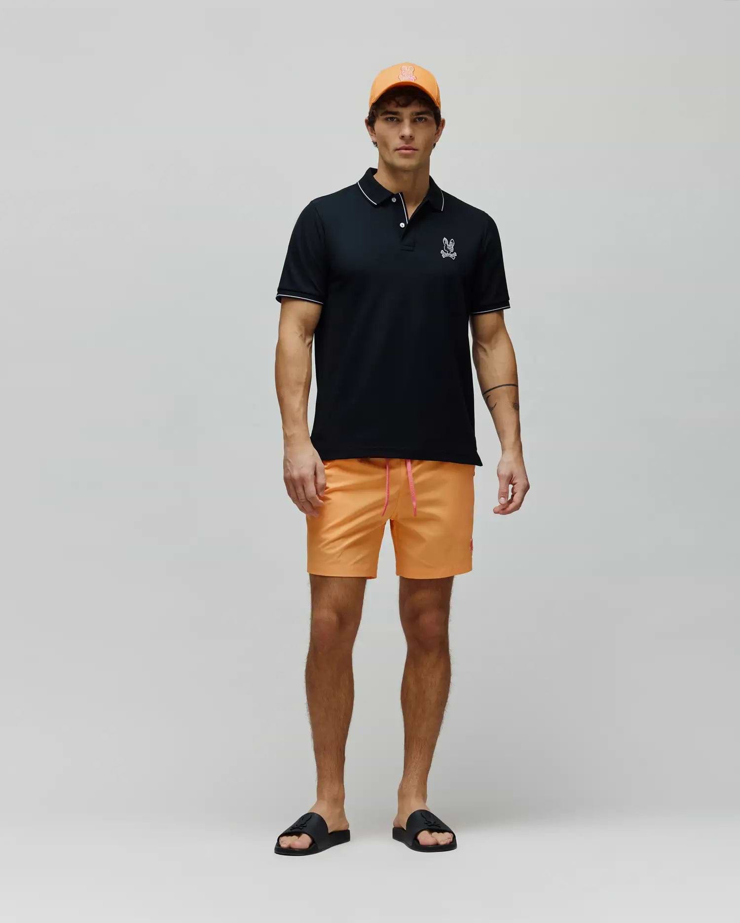 Mens Olivier Pique Polo in black with white trim and embroidered Psycho Bunny logo, paired with bright orange shorts featuring a pink drawstring and small bunny logo, an orange cap, and black logo slides. (4)