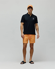 Mens Olivier Pique Polo in black with white trim and embroidered Psycho Bunny logo, paired with bright orange shorts featuring a pink drawstring and small bunny logo, an orange cap, and black logo slides. (4)