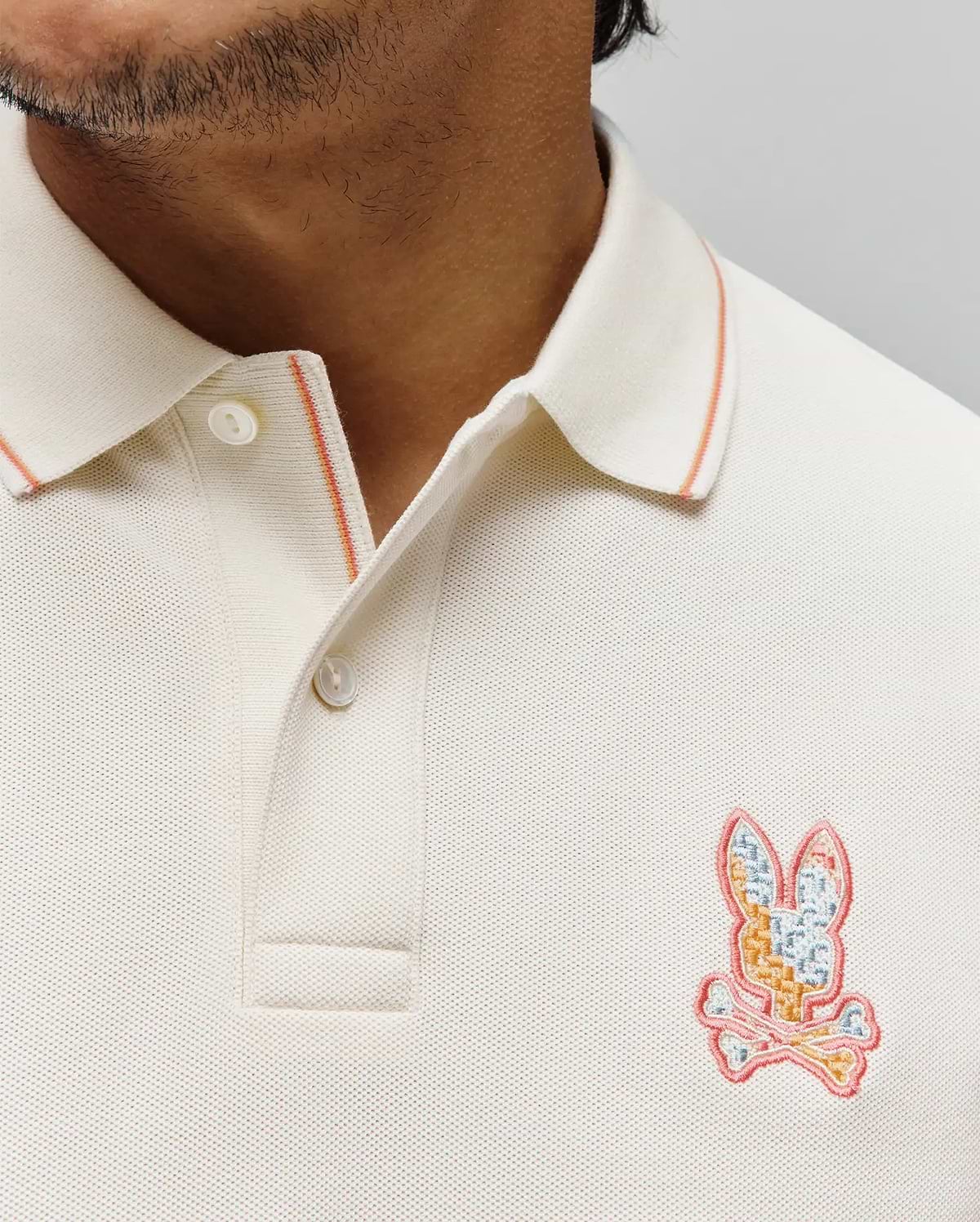 Mens Olivier Pique Polo in cream pique fabric, featuring a ribbed collar with orange tipping and a vibrant multi-color embroidered Psycho Bunny skull logo on the left chest. (2)