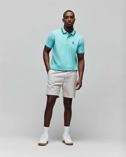 Mens Clark Pique Polo in aqua, classic fit, with dark blue and white striped collar and cuffs, and a small embroidered Psycho Bunny skull logo. Paired with light beige drawstring shorts. (3)
