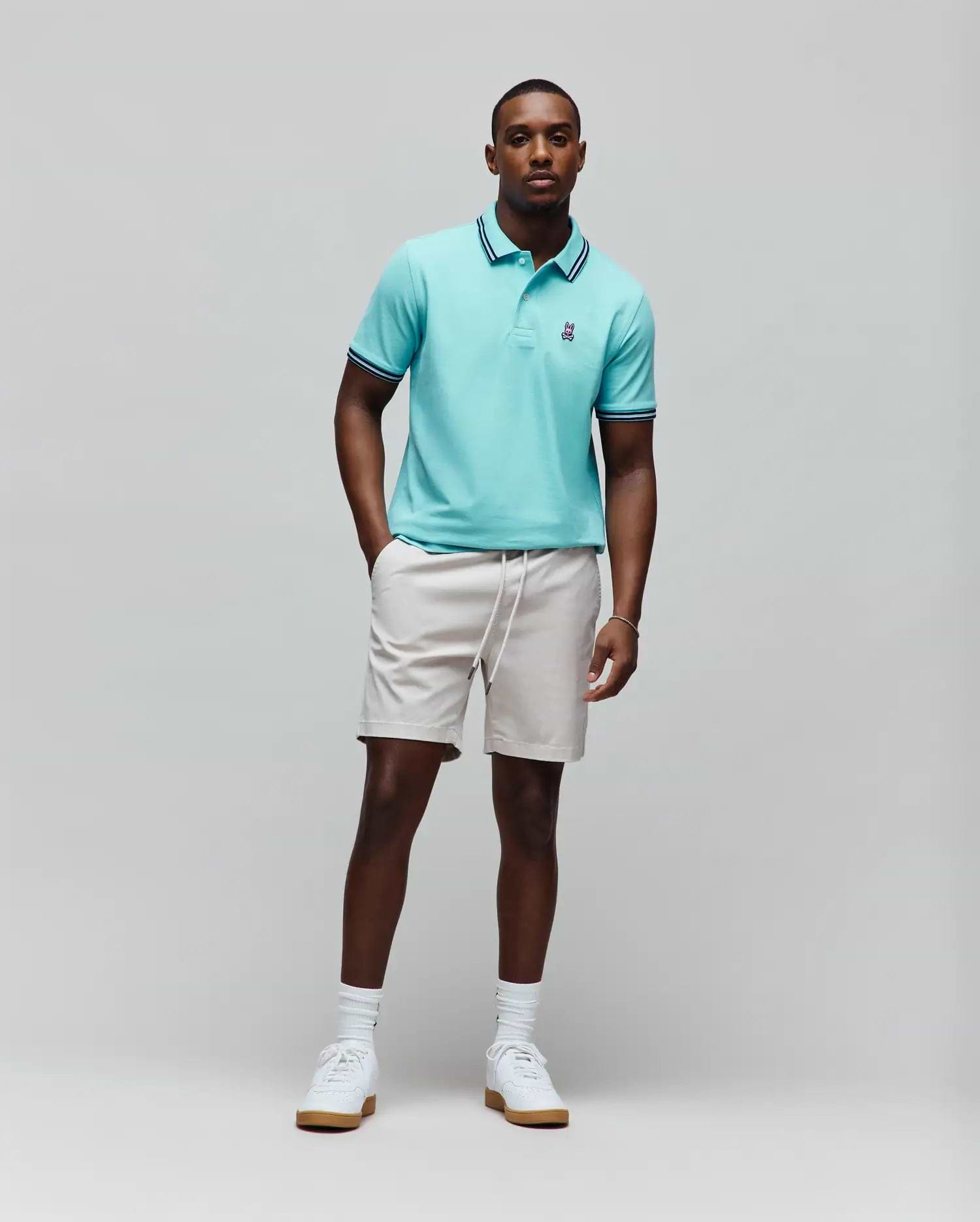 Mens Clark Pique Polo in aqua, classic fit, with dark blue and white striped collar and cuffs, and a small embroidered Psycho Bunny skull logo. Paired with light beige drawstring shorts. (3)