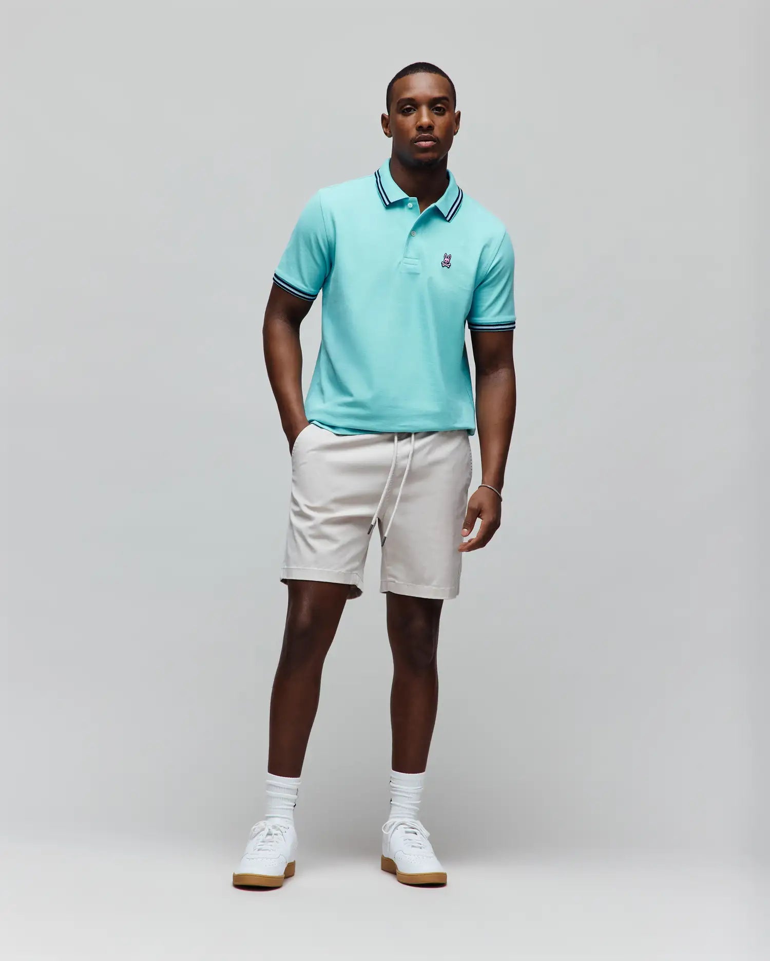 Mens Clark Pique Polo in aqua, classic fit, with dark blue and white striped collar and cuffs, and a small embroidered Psycho Bunny skull logo. Paired with light beige drawstring shorts. (3)