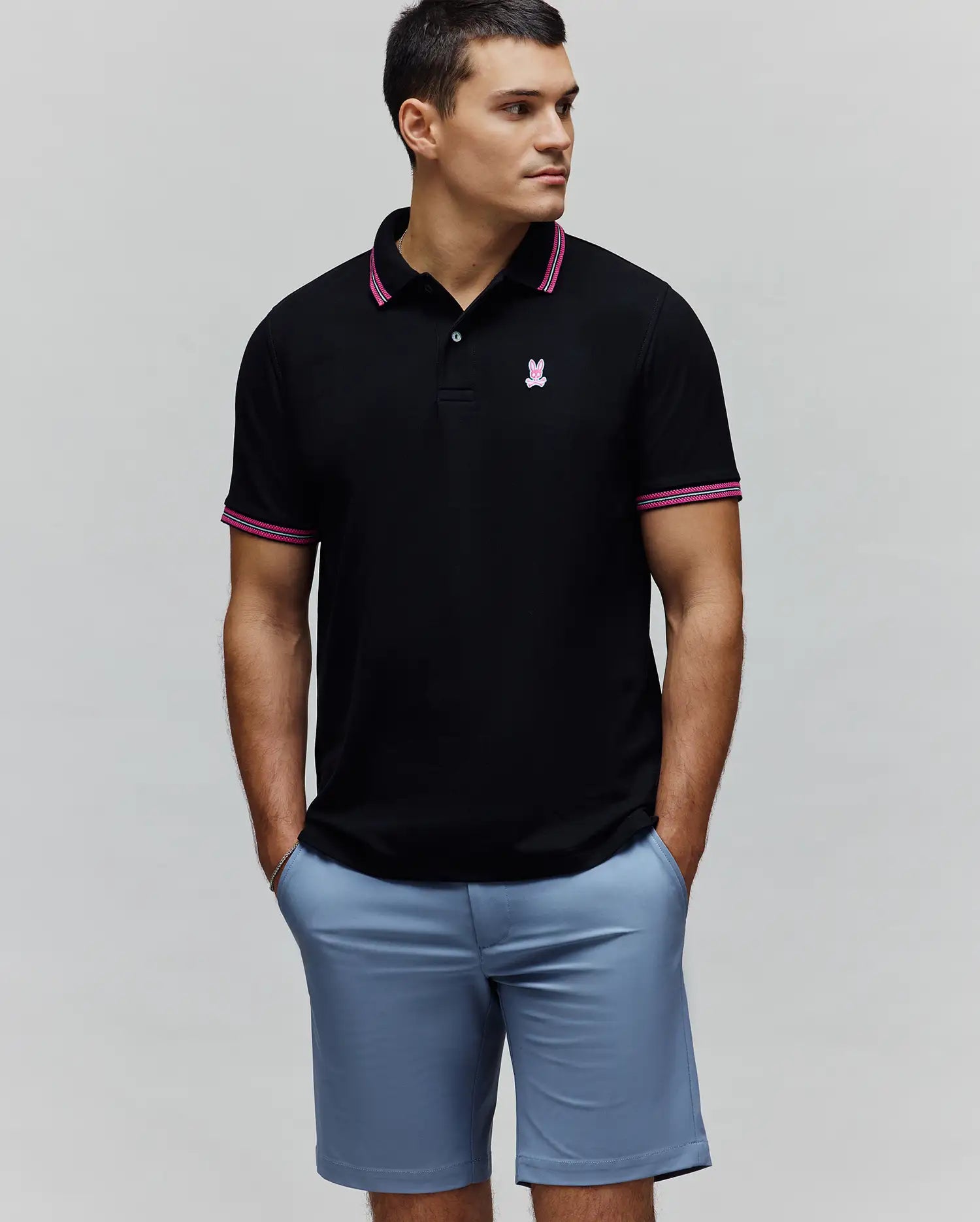Mens Clark Pique Polo. Black classic fit pique polo with a small embroidered pink Psycho Bunny skull logo on the left chest, accented by pink and white striped collar and sleeve cuffs.