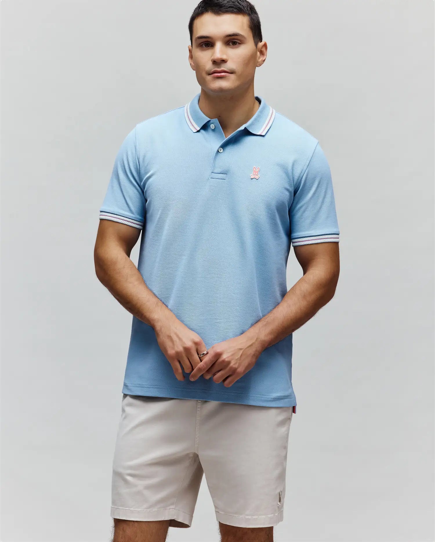 Mens Clark Pique Polo, a light blue classic fit pique polo with white and red striped collar and cuffs, featuring a small embroidered pink Psycho Bunny logo on the left chest. (3)
