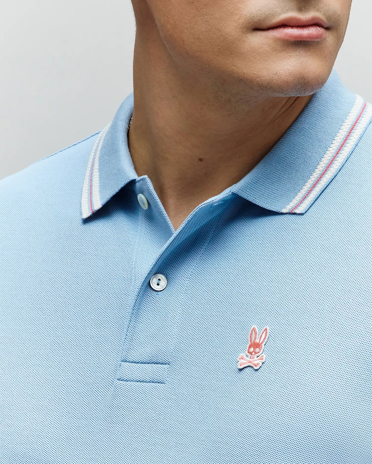 Mens Clark Pique Polo, a light blue classic fit pique polo featuring a white and red striped collar and a small embroidered pink Psycho Bunny skull logo on the left chest. (2)