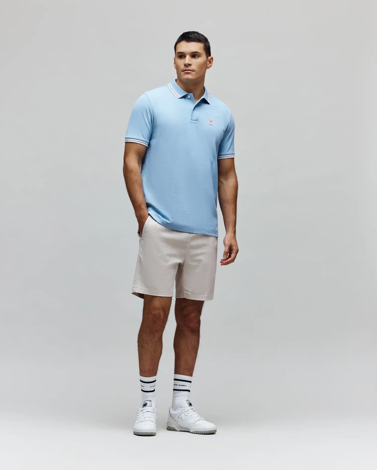 Mens Clark Pique Polo, a light blue classic fit pique polo with white and pink striped collar and cuffs, featuring a small embroidered pink Psycho Bunny skull logo on the left chest. (4)