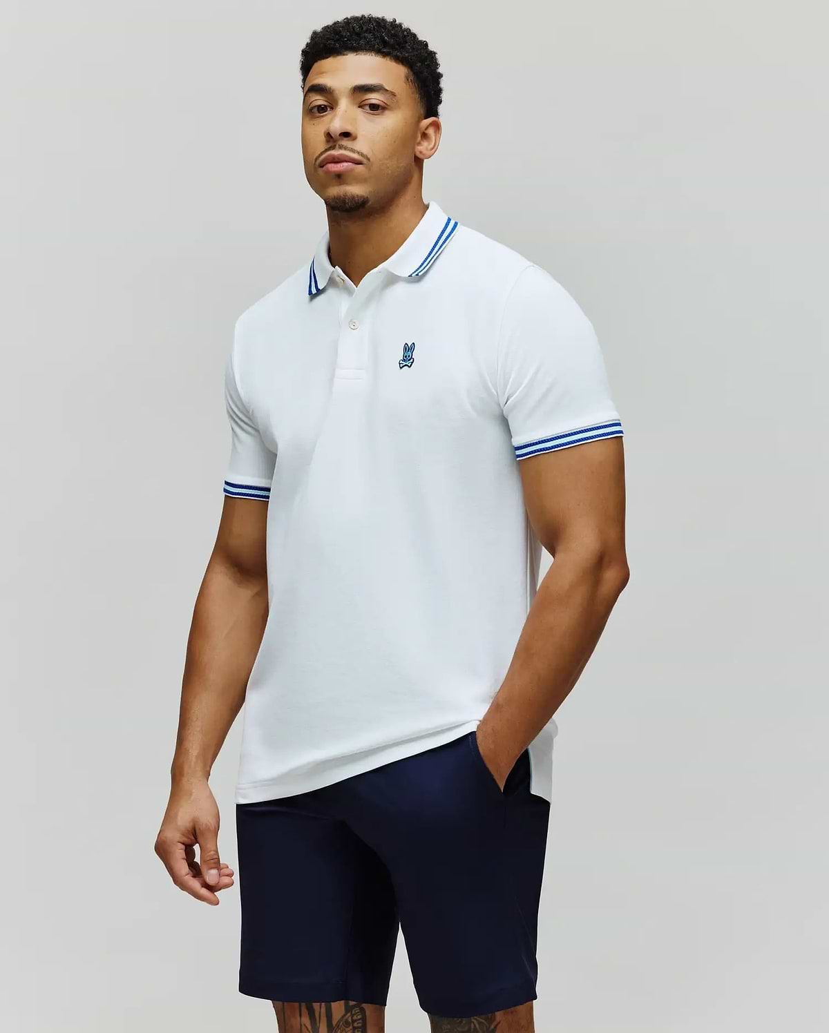 Mens Clark Pique Polo white classic fit pique polo with blue double-striped collar and cuffs, featuring a small embroidered blue Psycho Bunny skull logo on the left chest. (1)