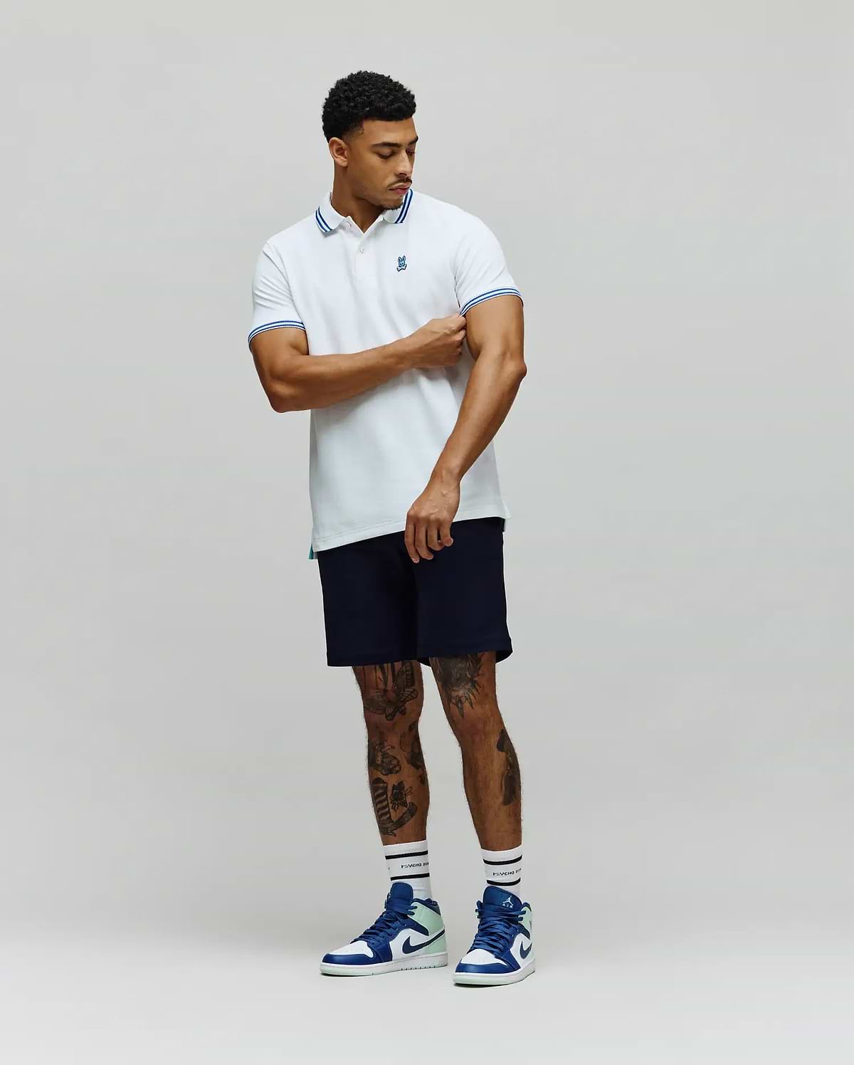 Mens Clark Pique Polo, a classic fit white pique polo shirt with blue double stripe accents on the collar and cuffs, featuring a small embroidered blue Psycho Bunny skull logo on the left chest. (4)