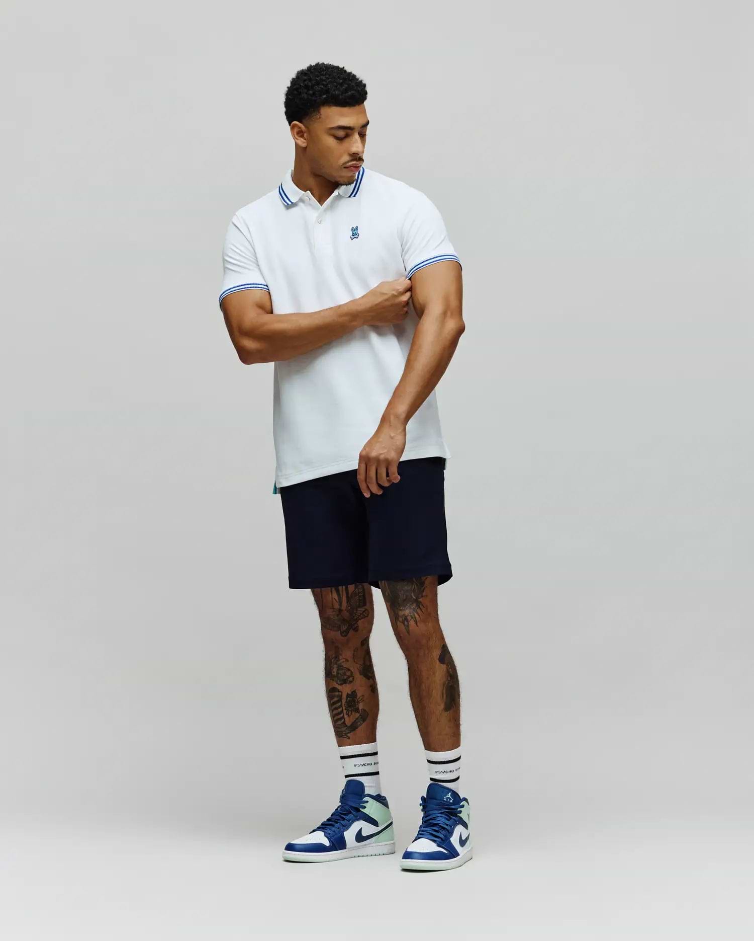 Mens Clark Pique Polo, a classic fit white pique polo shirt with blue double stripe accents on the collar and cuffs, featuring a small embroidered blue Psycho Bunny skull logo on the left chest. (4)