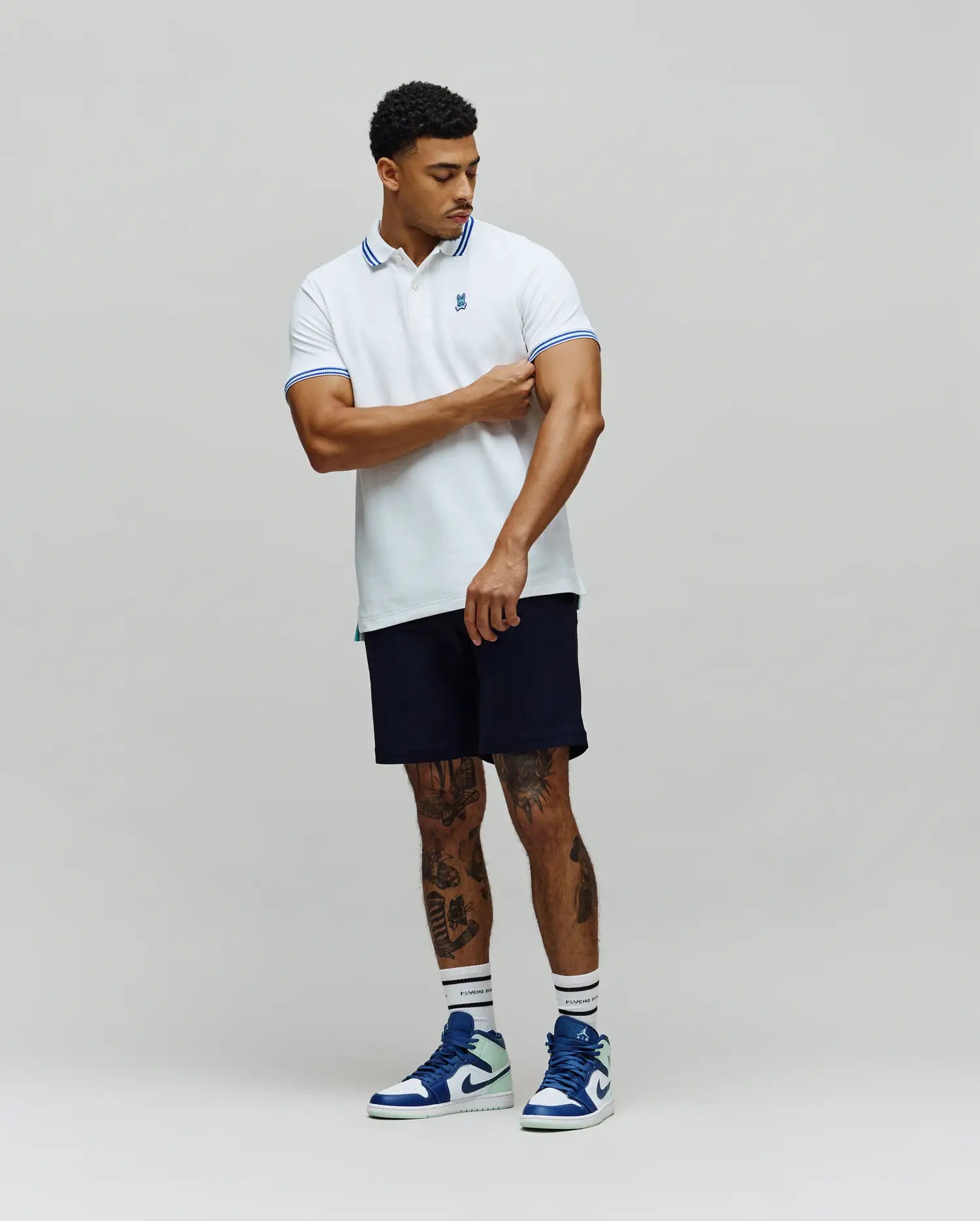 Mens Clark Pique Polo, a classic fit white pique polo shirt with blue double stripe accents on the collar and cuffs, featuring a small embroidered blue Psycho Bunny skull logo on the left chest. (4)