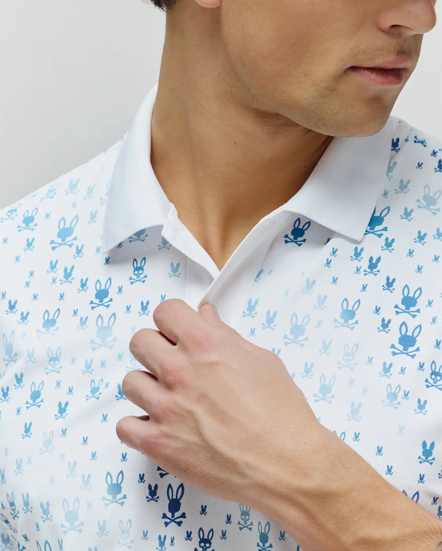 Mens Nev Sport Polo, white pique knit polo featuring an all-over gradient blue Psycho Bunny skull logo pattern in varying sizes, with a crisp white collar.