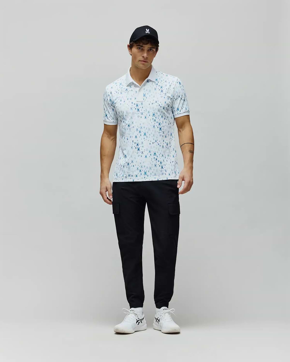 Mens Nev Sport Polo, white pique knit with an all-over light blue Psycho Bunny skull print, classic fit, featuring a small embroidered bunny logo on the left chest and ribbed collar/cuffs.