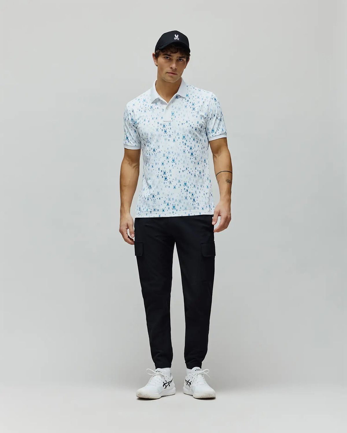 Mens Nev Sport Polo, white pique knit with an all-over light blue Psycho Bunny skull print, classic fit, featuring a small embroidered bunny logo on the left chest and ribbed collar/cuffs.