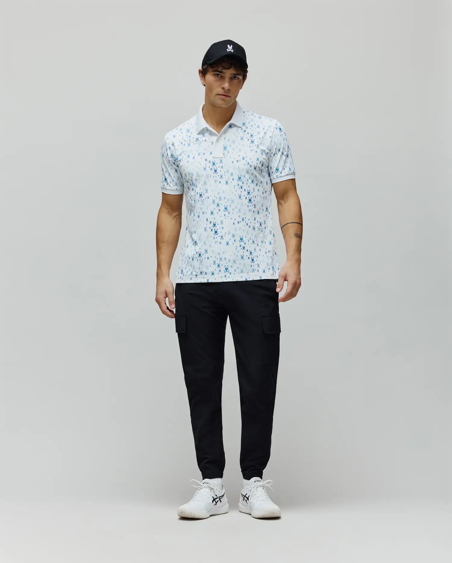 Mens Nev Sport Polo, white pique knit with an all-over light blue Psycho Bunny skull print, classic fit, featuring a small embroidered bunny logo on the left chest and ribbed collar/cuffs.