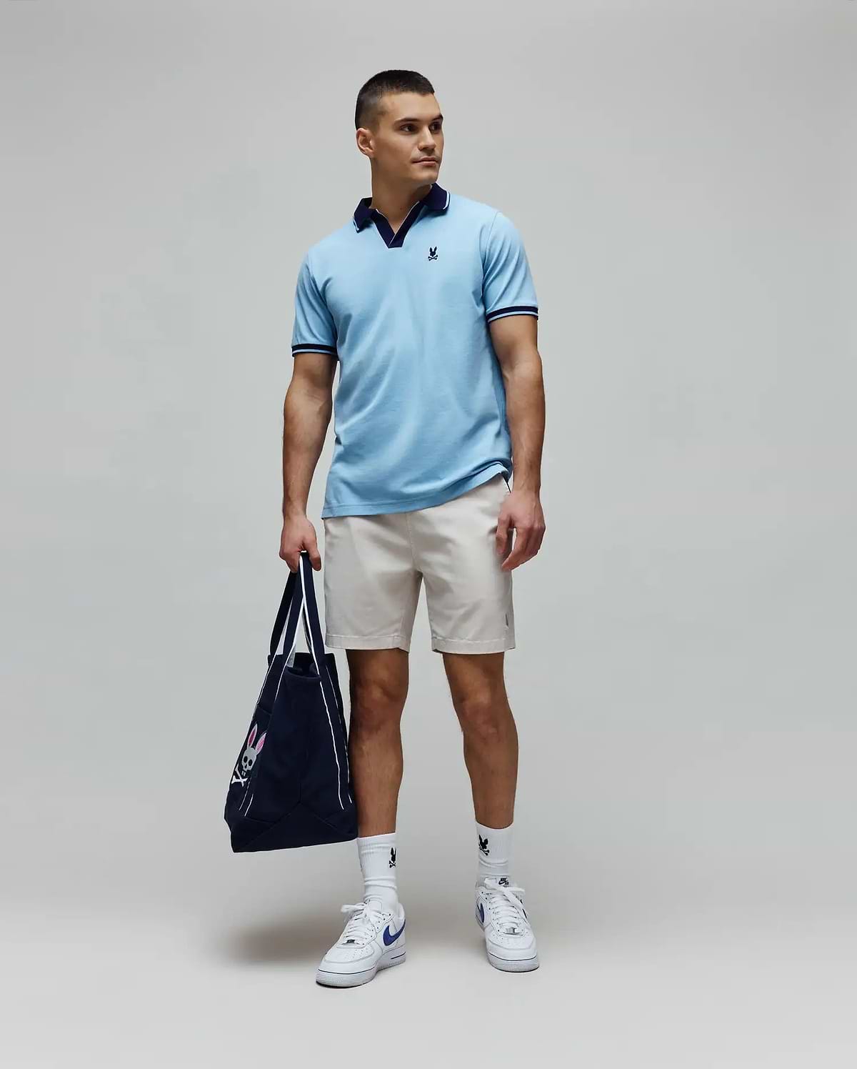 Mens Christiano Pique Polo in light blue, classic fit, with navy collar and cuffs, featuring an embroidered Psycho Bunny logo. Paired with beige shorts, white logo socks, and a navy Psycho Bunny tote bag. (3)