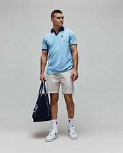 Mens Christiano Pique Polo in light blue, classic fit, with navy collar and cuffs, featuring an embroidered Psycho Bunny logo. Paired with beige shorts, white logo socks, and a navy Psycho Bunny tote bag. (3)
