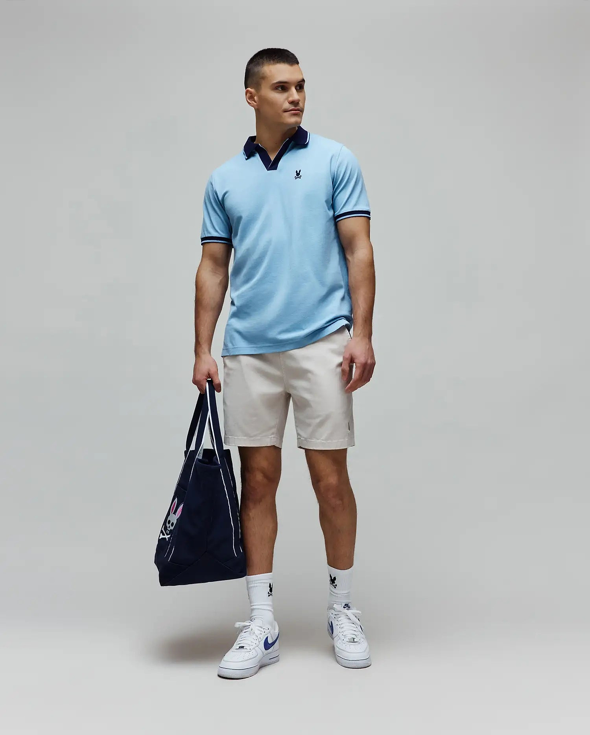 Mens Christiano Pique Polo in light blue, classic fit, with navy collar and cuffs, featuring an embroidered Psycho Bunny logo. Paired with beige shorts, white logo socks, and a navy Psycho Bunny tote bag. (3)
